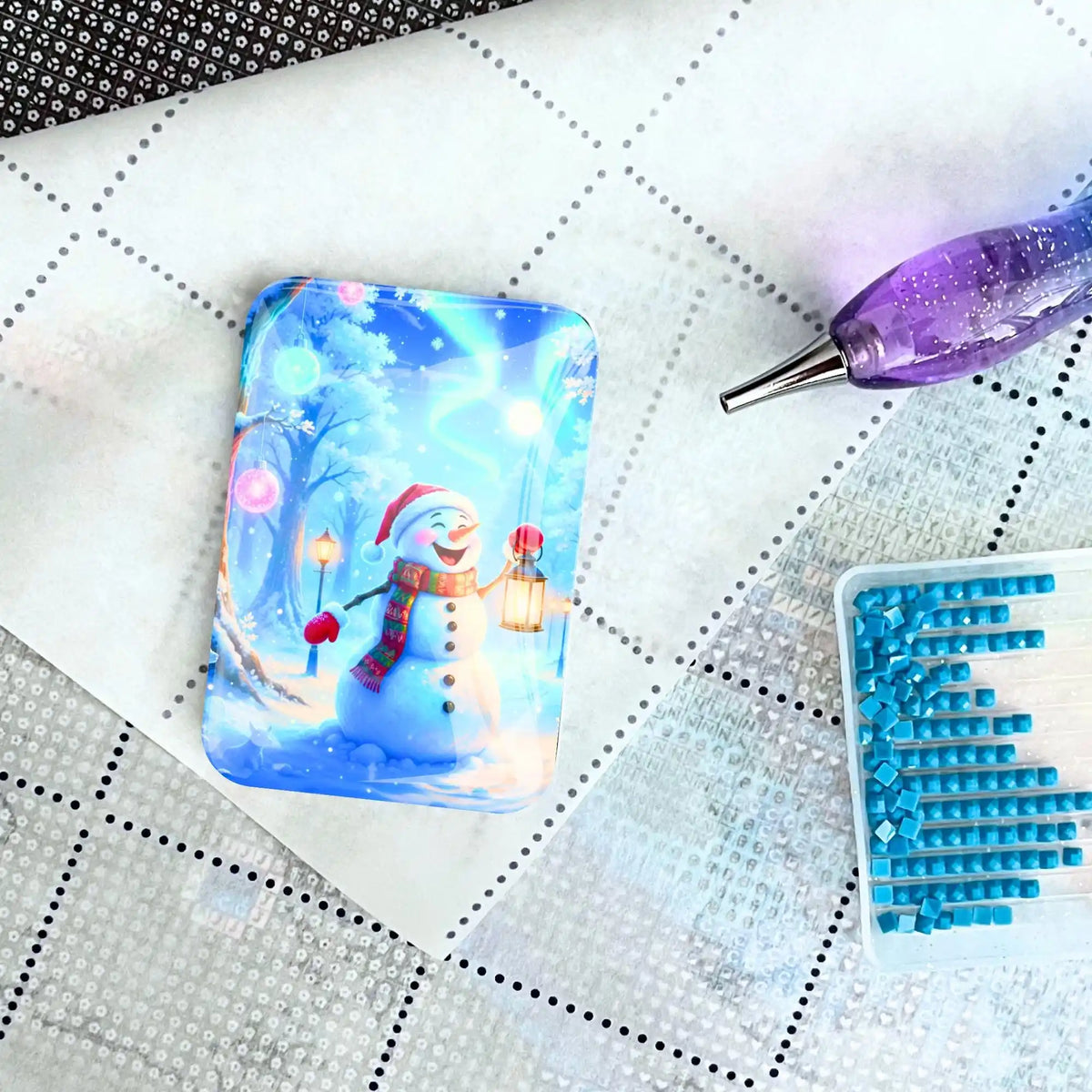 Diamond Painting Glas Cover Minder mit Schneemann auf Leinwand mit lila Stift und blauen Steinen.