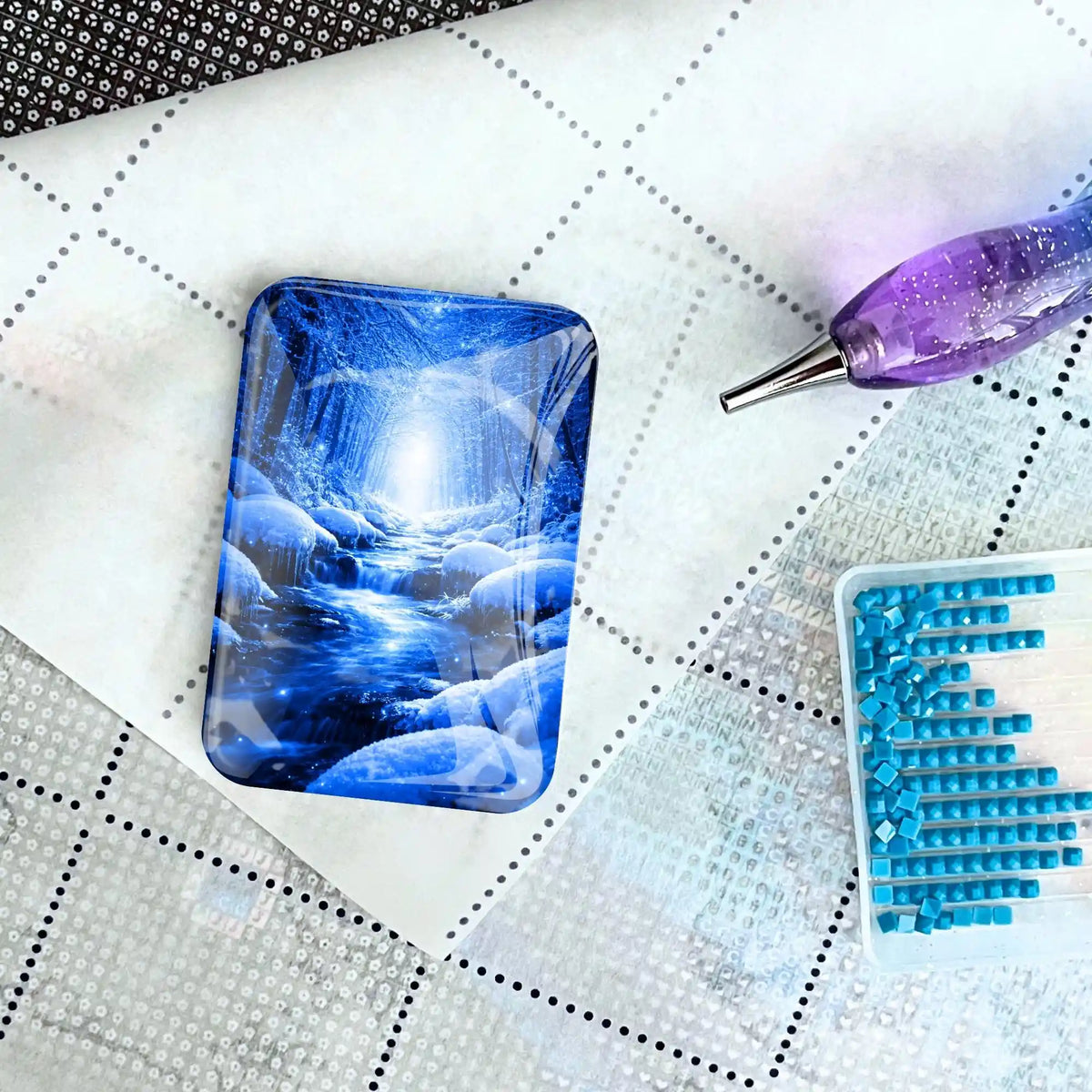 Diamond Painting Glas Cover Minder mit blauem Winterwald-Motiv auf Leinwand neben Schiffchen und Glitzer-Stift